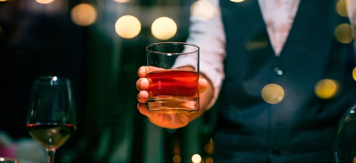 glass of whiskey in a mans hand over one ice cube standing in a bar